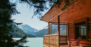 Home Tranquil scene of a wooden cabin overlooking a serene mountain lake, surrounded by lush forest.