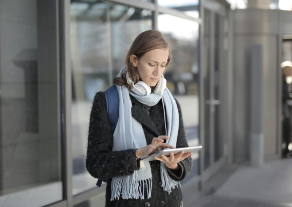 Young woman stands outside using a tablet, wearing headphones and a scarf, representing technology and travel.